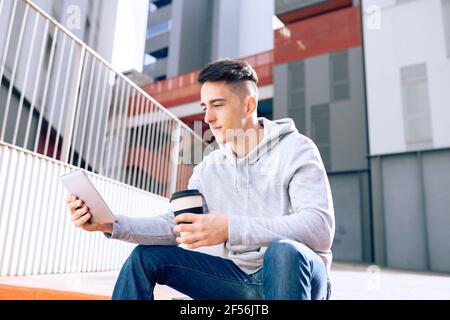 Young man holding reusable coffee cup while using digital tablet against building Stock Photo