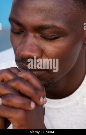 Close up portrait of young african man praying with hands clasped and eyes closed Stock Photo