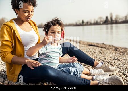 Boy blowing in bubble wand while sitting with woman on pebble Stock Photo