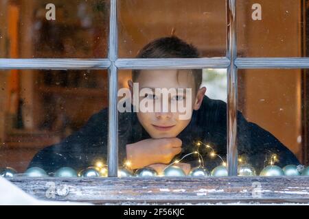 Boy with hand on chin looking through window while sitting at home during Christmas Stock Photo