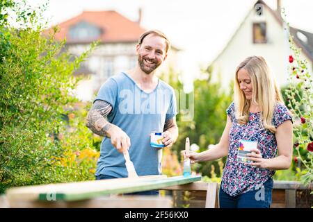 Mature man smiling while painting wooden plank in garden with girlfriend Stock Photo