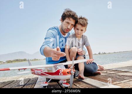 Father talking over airplane toy with son while sitting on pier Stock Photo