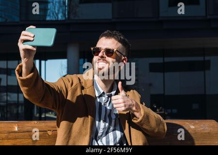 Smiling businessman gesturing thumbs while taking selfie through mobile phone sitting on bench Stock Photo