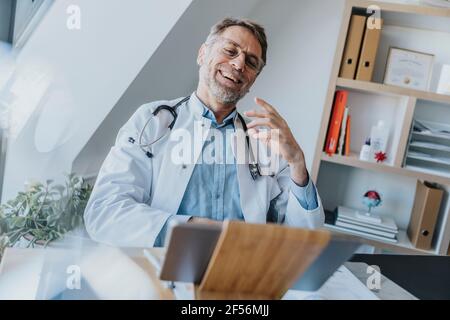 Male healthcare worker talking on video call with patient over digital tablet while sitting at doctor's office Stock Photo