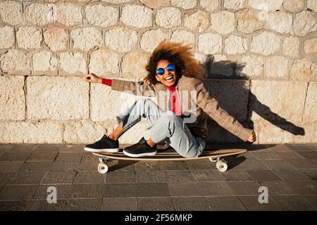 Cheerful woman with arms outstretched sitting on skateboard against wall during sunny day Stock Photo