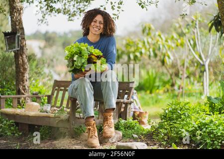 Smiling woman holding leafy vegetables while sitting on wooden bench in garden Stock Photo