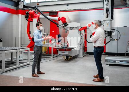 Young engineer discussing with mature man by robotics in factory Stock Photo