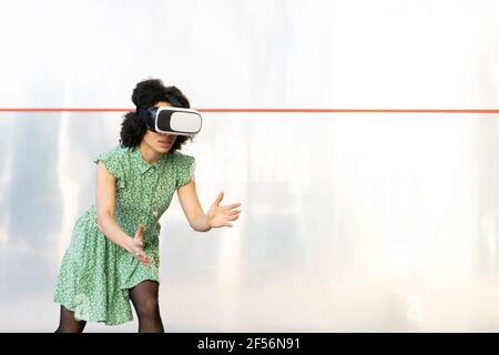 Young woman playing while wearing Virtual reality headset against wall Stock Photo