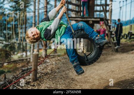 Playful boy swinging on tire swing during rope course in forest Stock Photo
