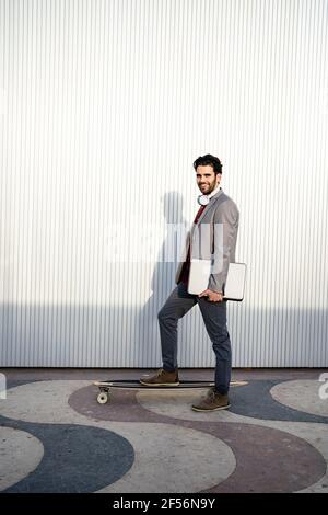 Smiling businessman with laptop standing on longboard by wall Stock Photo