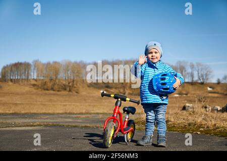 Cute boy with cycling helmet waving hand while standing by balance bike on road Stock Photo