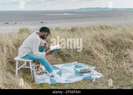 Concentrated young man reading book while sitting on stool amidst dried plants at beach Stock Photo