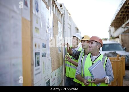 Three male construction workers inspecting bulletin board at construction site Stock Photo