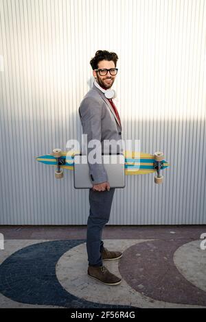 Smiling businessman with longboard and laptop standing on footpath by wall Stock Photo