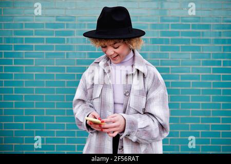 Happy curly haired woman in hat using mobile phone against wall Stock Photo