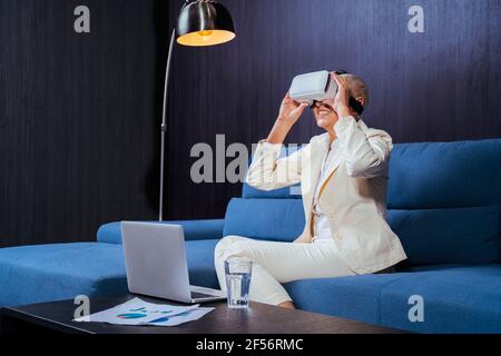 Smiling businesswoman using virtual reality headset while sitting by laptop on sofa Stock Photo