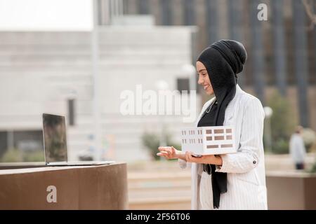 Young female architect holding architectural model during video call on laptop Stock Photo