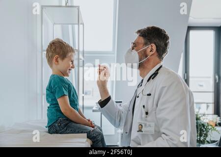 Pediatrician wearing protective face mask taking nasal swab test of boy while standing at clinic Stock Photo