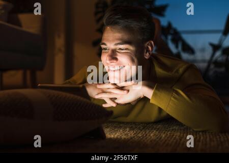 Smiling young man lying on carpet while watching videos through digital tablet in living room during night Stock Photo