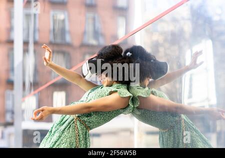Mixed race woman playing wearing Virtual reality headset while leaning against glass wall Stock Photo