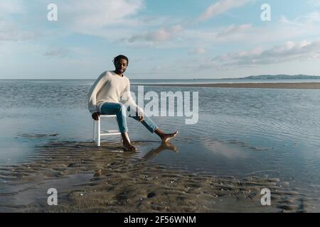 Smiling young man sitting on white stool at beach during sunset Stock Photo