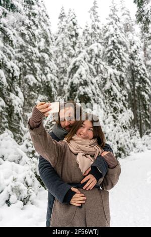 Girlfriend taking selfie through mobile phone with man embracing her from behind while standing at forest Stock Photo