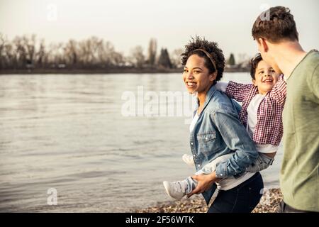 Smiling mother piggybacking son while walking with father by lake Stock Photo