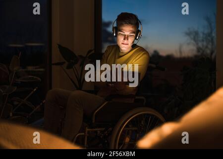 Young man in wheelchair wearing wireless headphones while using digital tablet sitting on wheelchair in living room Stock Photo