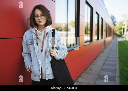 Young stylish queer girl, college student leaning on wall, holding tote-bag, smiling looking at camera, walking outdoors after classes finished Stock Photo