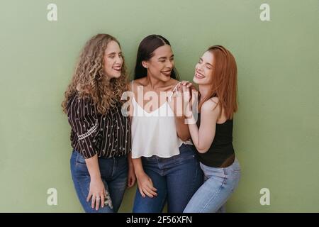 Cheerful young female friends standing against green wall Stock Photo
