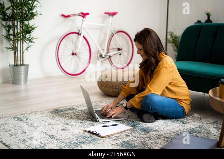 Smiling woman with cross legs sitting on floor while using laptop in living room Stock Photo