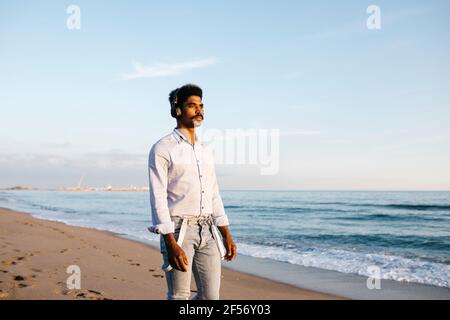 Handsome Afro man standing at beach listening music against sky Stock Photo