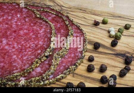 Makro closeup of isolated slices french fresh salami with mixed pepper crust and peppercorns on olive wood cutting board Stock Photo