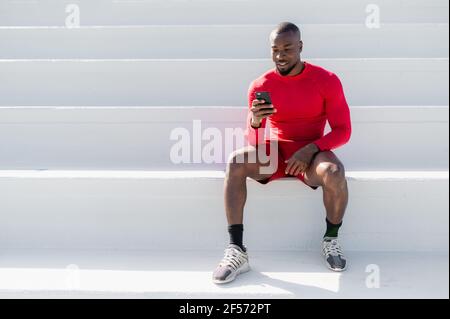 black African man with sportswear sitting on white steps using smartphone. Minimalism and copy space. Stock Photo
