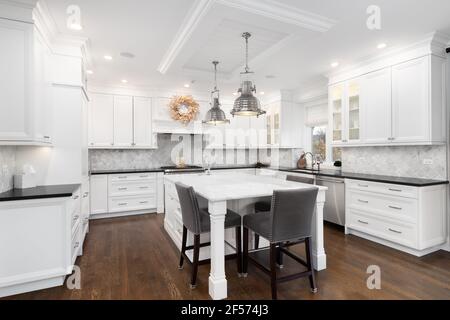 A modern white kitchen with chairs sitting at the large island, chrome lights hanging from the ceiling, and hardwood floors throughout. Stock Photo