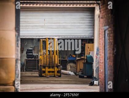 Old yellow fork lift truck parked in garage with roller shutter door down.  Undergoing repair and maintenance in old workshop warehouse. Stock Photo