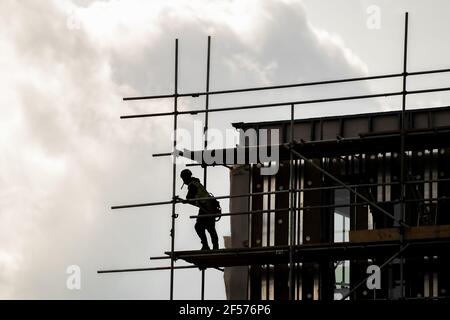 Silhouette scaffolder in PPE erecting framework of wooden planks and tall scaffolding poles high up on modern new building construction site. Looking Stock Photo