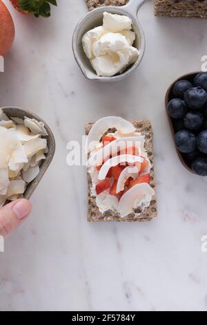 A healthy isolated cracker with coconut slices and pieces of strawberry in the middle surrounded by trays full of fruit and a bowl with cream cheese o Stock Photo
