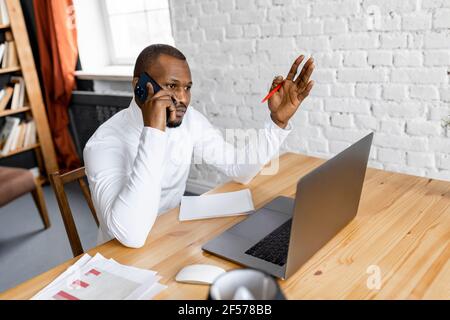 Serious african american male employee is sitting at the desk, having a phone conversation. Focused businessman discussing new project with colleagues Stock Photo