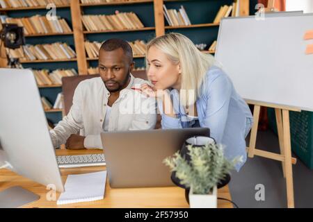 Serious multiethnic coworkers using computer, searching ideas for project. Young adult employees working together in modern office, developing new Stock Photo