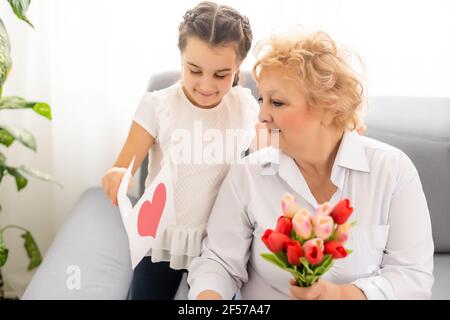 Sincere cute little granddaughter strong cuddling and kissing in cheek her 60s grandmother gave her pretty spring flowers congratulates with birthday Stock Photo