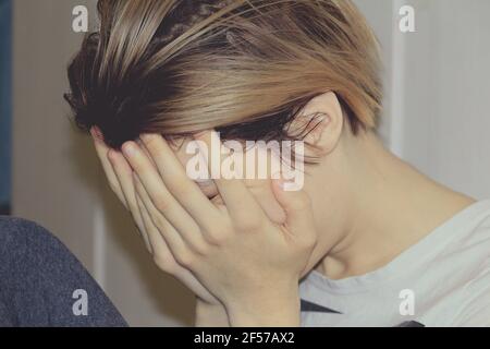 The girl covers her face with her hands. A young girl with blond hair sits in profile on the floor in a room and covers her face with her hands. Stock Photo