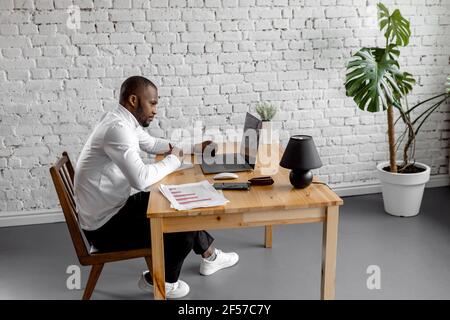 Focused african american male employee or student is using laptop, having online conference or lecture. Serious businessman is sitting at the desk in Stock Photo