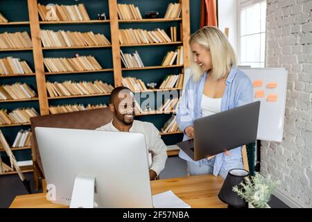 Happy friendly coworkers using laptop, smiling, working together in modern office. Successful business partners developing new business strategy or Stock Photo