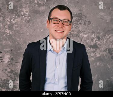 Close-up portrait of confident young successful caucasian businessman wearing a suit and glasses smiling looking into the camera against a concrete gr Stock Photo