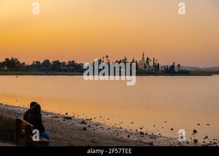 A romantic couple enjoys the sunset seated on a bench at Larnaca salt lake with the Hala Sultan Tekke mosque in the background Stock Photo