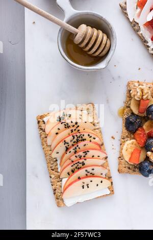 A delicious cracker with apple slices topped with black sesame grains on cream cheese next to a bowl full of honey with a wooden dipper inside. Stock Photo