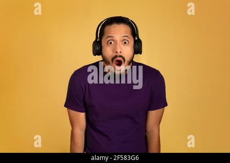 Young man with surprised face listening to music using headphones. Yellow background. Stock Photo
