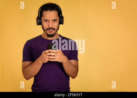 Young man listening music using headphones and playing with the smartphone. Yellow background. Stock Photo