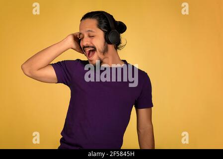 Young man smiling and singing while listening to music using headphones and playing with the smartphone. Yellow background. Stock Photo